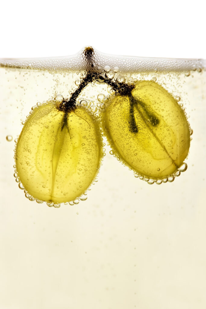 Deux grains de raisin Chardonnay plongé dans une coupe de champagne. On note la transparence des grains et l’effervescence à la surface. / Two Chardonnay grapes plunged into a glass of champagne. Note the transparency of the grapes and the effervescence on the surface.
