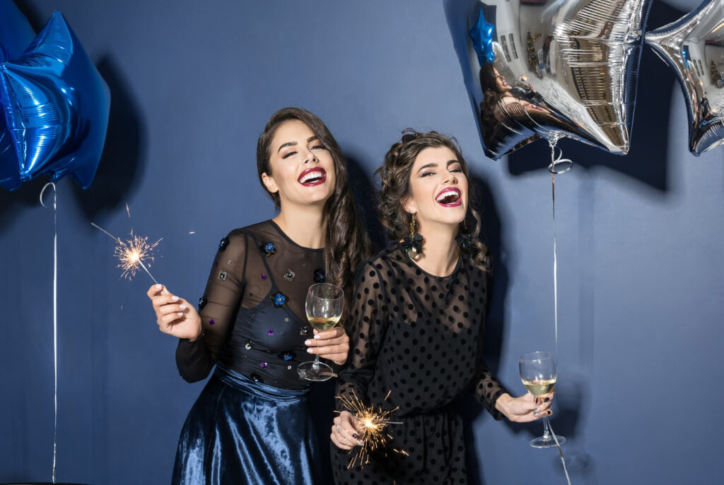 deux femmes verre à la main rient aux éclats et tiennent des ballons bleus et argentés en forme d’étoiles, ambiance festive. Elles tiennent chacune une coupe de champagne / Two women holding glasses, laughing and holding blue and silver balloons in the shape of stars, create a festive atmosphere. They are each holding a glass of champagne