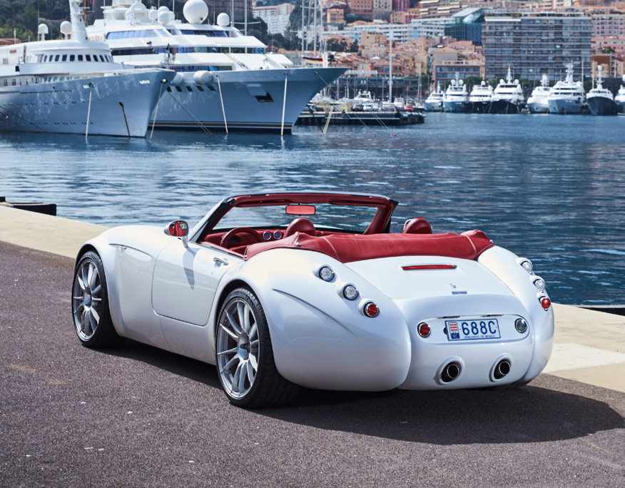 Voiture sport garée sur le port en bord de mer, yachts sur la mer en arrière-plan. La voiture est grise et décapotable, son intérieur est rouge / Sports car parked on the seafront harbour, yachts on the sea in the background. The car is grey and convertible, with a red interior