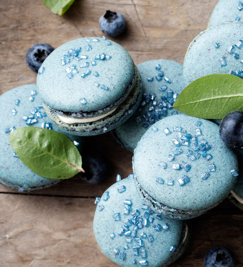 Macarons bleus posés sur une table en bois, fourrés à la vanille. On aperçoit deux feuilles de menthe et des myrtilles en décorations. / Blue macaroons with vanilla filling on a wooden table. Two mint leaves and blueberries can be seen as decorations.