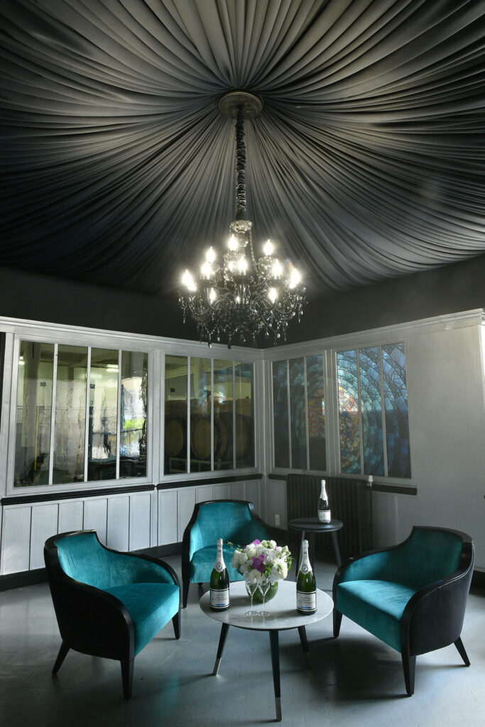 Petit salon au Champagne Charles de Cazanove avec 3 fauteuils bleu canard, une table basse. Un bouquet est posé sur la table basse, un lustre se trouve en suspension au-dessus de celle-ci. / Small lounge in Champagne Charles de Cazanove with 3 duck-blue armchairs and a coffee table. A bouquet sits on the coffee table and a chandelier hangs above it.