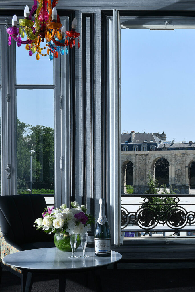 Intérieur du Champagne Charles de Cazanove, un magnum de Tête de Cuvée est posé sur la table basse avec un bouquet de fleurs. Une fenêtre laisse apparaître la Porte de Mars ensoleillé. / Inside Champagne Charles de Cazanove, a magnum of Tête de Cuvée sits on the coffee table with a bouquet of flowers. A window reveals the sunny Porte de Mars.