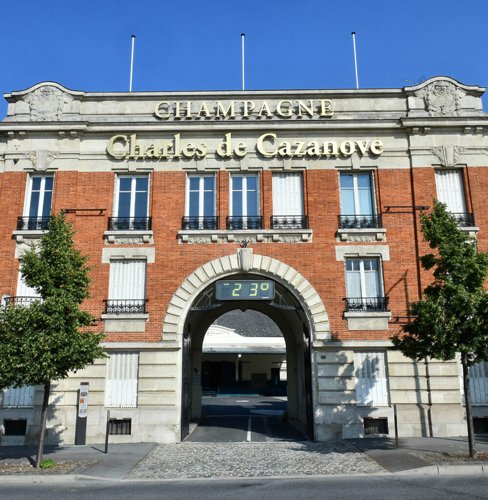 Façade en brique ensoleillée du Champagne Charles de Cazanove avec l’enseigne mise en évidence et son grand porche d’entrée. / The sunny brick façade of Champagne Charles de Cazanove, with its prominent sign and large entrance porch.
