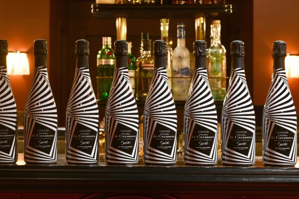 Rangée de huit bouteilles de Cuvée Senses aux rayures noires et blanches alignées et posées sur un bar en bois avec, au fond, des bouteilles d’alcool. / A row of eight bottles of Cuvée Senses with black and white stripes lined up on a wooden bar with bottles of spirits at the back.
