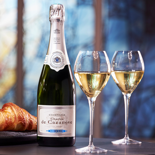 Bouteille de champagne Blanc de Blancs Charles de Cazanove avec deux coupes, le tout posé sur une table avec un morceau de croissant et des vitraux en arrière-plan. / Bottle of Charles de Cazanove Blanc de Blancs champagne with two glasses, all placed on a table with a piece of croissant and stained glass windows in the background.