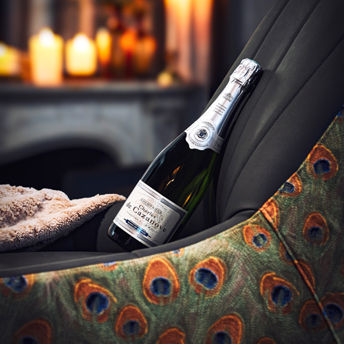 Bouteille de Blanc de Blancs couchée sur un fauteuil aux motifs animaux de paon, cheminée en arrière-plan et bougies allumée. Ambiance chaleureuse. / Bottle of Blanc de Blanc lying on an armchair with peacock animal motifs, fireplace in the background and candles lit. A warm atmosphere.