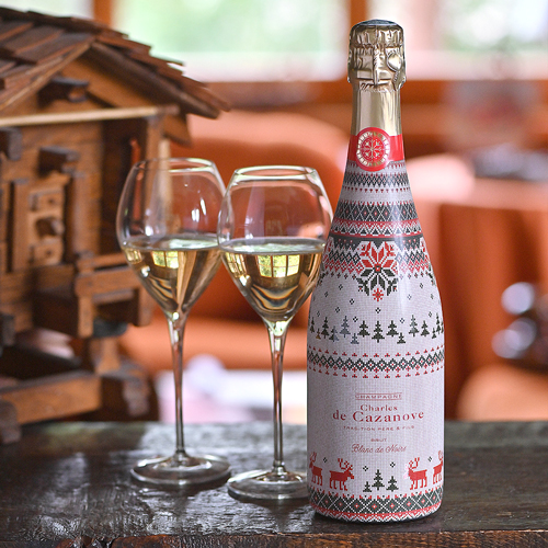 Bouteille de champagne Charles de Cazanove Happy Holidays 2 aux décors de pull de Noël dans une ambiance chalet en bois, deux flûtes de champagne à côté de la bouteille / Bottle of Charles de Cazanove Happy Holidays 2 champagne with Christmas jumper decorations in a wooden chalet atmosphere, two champagne flutes next to the bottle.