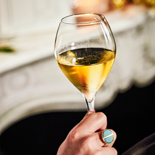 Coupe de champagne en zoom tenue par une main avec une bague. En fond une cheminée. / Zooming champagne glass held by a hand with a ring. In the background, a fireplace.