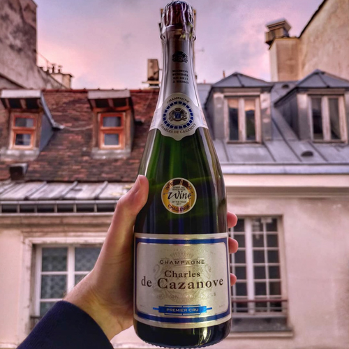 Bouteille de champagne Charles de Cazanove Premier Cru tenue par une main, façade en arrière-plan. / Bottle of Charles de Cazanove Premier Cru champagne held in one hand, facade in background.
