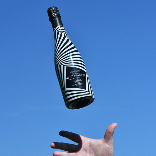 Bouteille de champagne Charles de Cazanove Senses lancée par une main sur un fond de ciel bleu. Bouteille rayée noire et blanches / Charles de Cazanove Senses champagne bottle thrown by a hand against a blue sky. Black and white striped bottle