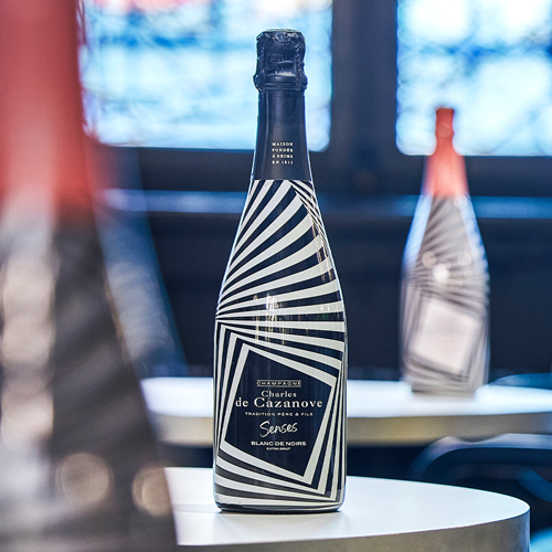 Bouteille de Senses posée sur une table, en arrière-plan et floutée apparaît la version de la bouteille en version rosée (coiffe rose) / Bottle of Senses placed on a table, with the pink version of the bottle (pink cap) in the background and out of focus.