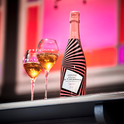 Bouteille de Senses Rosé Champagne Charles de Cazanove posée sur un bar à côté de deux flûtes, avec en fond des vitraux de couleur rose. Bouteille à rayures noire et rose / Bottle of Senses Rosé Champagne Charles de Cazanove placed on a bar next to two flutes, with pink stained glass windows in the background. Bottle with black and pink stripes