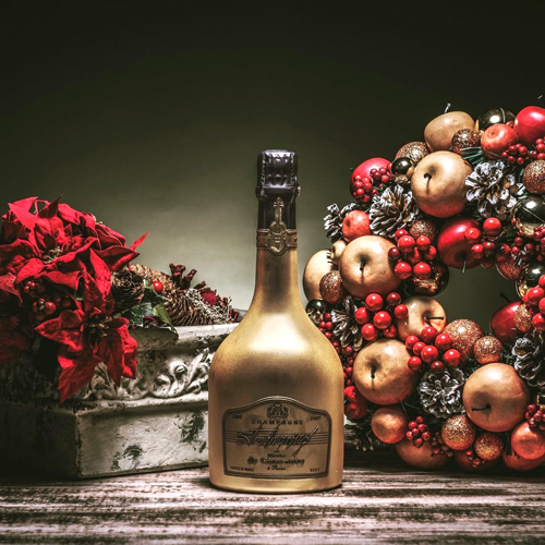 Bouteille de champagne Charles de Cazanove Stradivarius peinte en or avec une couronne de Noël en fond d’image / Charles de Cazanove Stradivarius champagne bottle painted gold with a Christmas wreath in the background