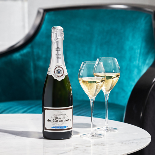 Bouteille de champagne Tête de cuvée Charles de Cazanove posée sur une table basse en marbre avec deux coupes de champagne. Fauteuil bleu canard et noir en fond d’image. / Bottle of champagne Tête de cuvée Charles de Cazanove on a marble coffee table with two glasses of champagne. Duck blue and black armchair in the background.