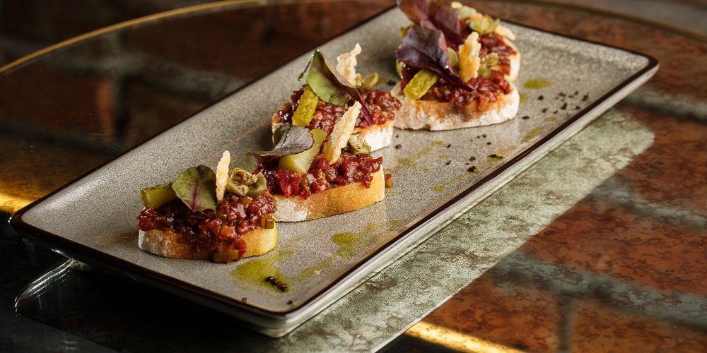 Tartare de bœuf sur toast de pain de campagne parsemé d’huile d’olive et de chips de légumes dans un plat rectangulaire / Beef tartare on country bread toast sprinkled with olive oil and vegetable chips in a rectangular dish.