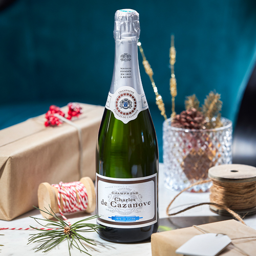 Préparatifs de Noël avec un bouteille de Tête de cuvée, cadeaux et décorations posés sur une table autour de la bouteille / Christmas preparations with a bottle of Tête de cuvée, gifts and decorations placed on a table around the bottle.