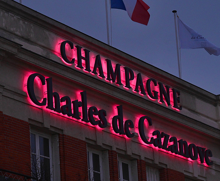 Gros plan sur l’enseigne Charles de Cazanove qui se trouve sur la façade du bâtiment. L’enseigne est éclairée de rosé, trois drapeaux se trouvent sur la haut de la façade, on aperçoit le drapeau de la France. / Close-up of the Charles de Cazanove sign on the façade of the building. The sign is illuminated in pink, and three flags can be seen at the top of the façade, including the flag of France.