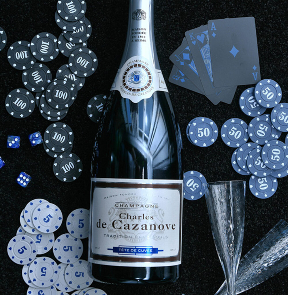 Magnum de Tête de Cuvée couché sur de la moquette noire. Autour se trouvent des jetons de poker, des cartes de jeu ainsi que deux flûtes de champagne en cristal. / Magnum of Tête de Cuvée lying on black carpet. Around it are poker chips, playing cards and two crystal champagne flutes.
